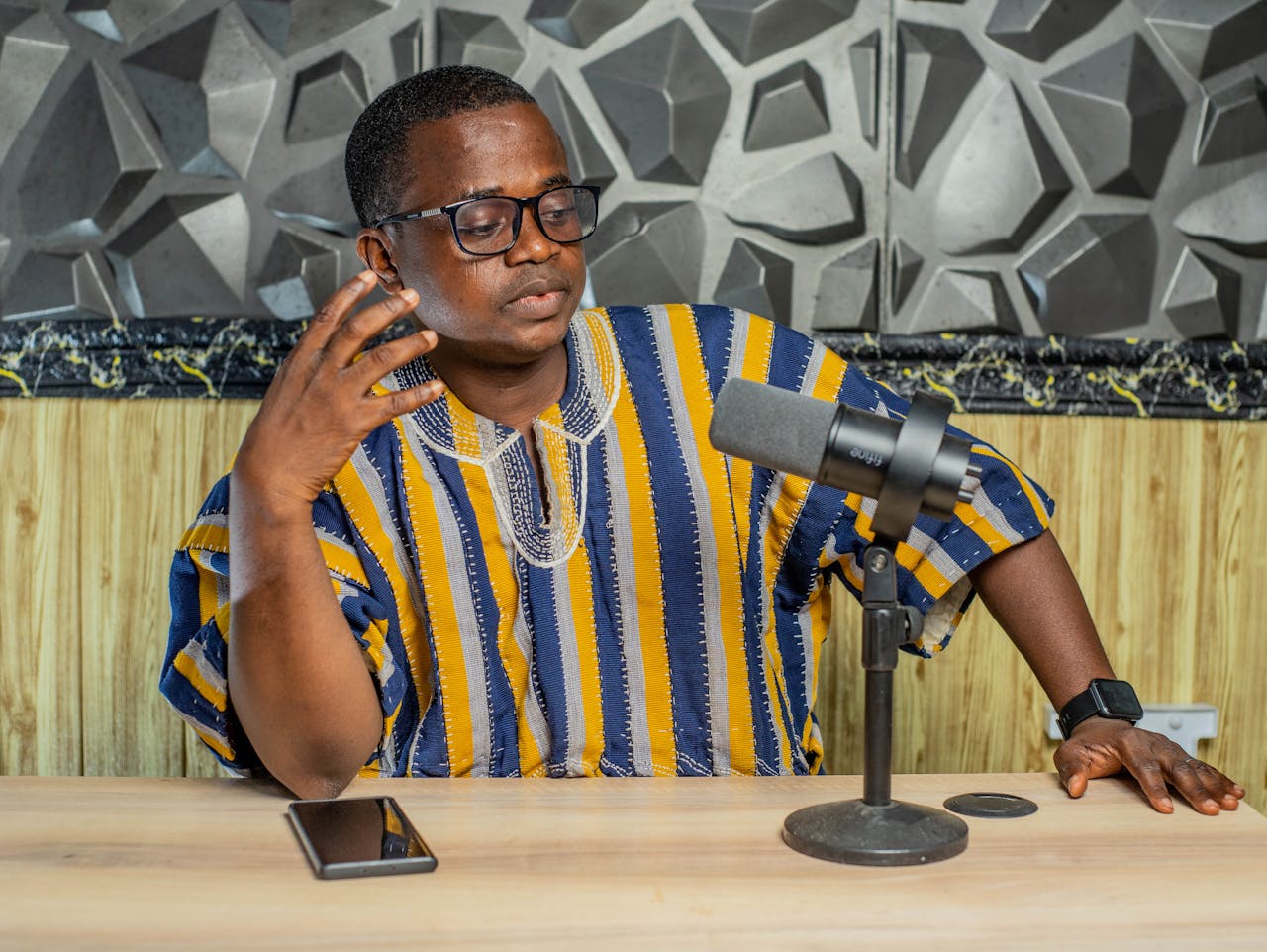 An adult man in a colorful Kente shirt recording a podcast. Indoor setting with microphone and smartphone.